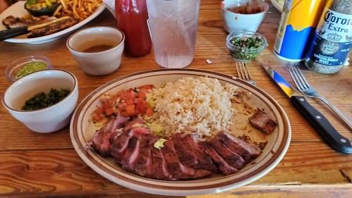 A plate of sliced steak with rice, diced tomatoes, and sauces, accompanied by drinks on a wooden table.