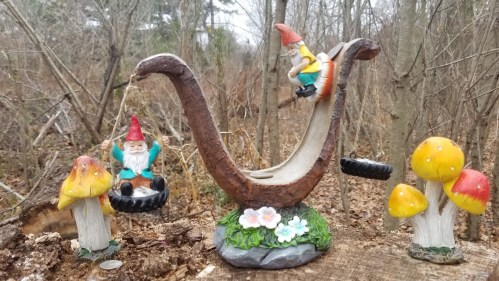 A whimsical scene featuring gnome figurines, a slide, and colorful mushrooms in a wooded area.