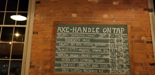 Chalkboard menu listing various beers on tap, with names, styles, and prices, set against a brick wall.