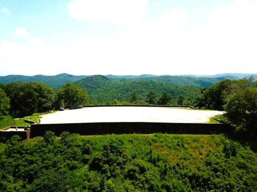 A wide, empty parking lot surrounded by lush green hills under a bright blue sky.
