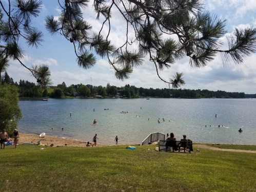 A sunny lakeside scene with people swimming, a sandy beach, and trees framing the view.