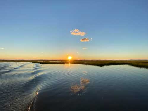 Sunset over calm waters, reflecting the sky and clouds, with grassy shoreline in the background.