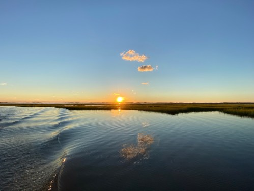 Sunset over calm waters, reflecting the sky and clouds, with grassy shoreline in the background.