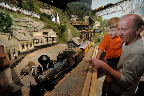 A man and a boy observe a detailed model train set, pointing at the miniature train and surrounding scenery.