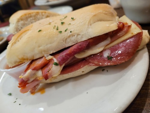 A close-up of a sandwich with layers of meats and cheese, served on a soft roll, garnished with herbs.
