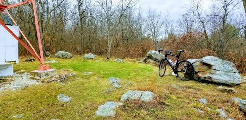 A black bicycle leans against a large rock near a tower in a wooded area with sparse trees and overcast skies.