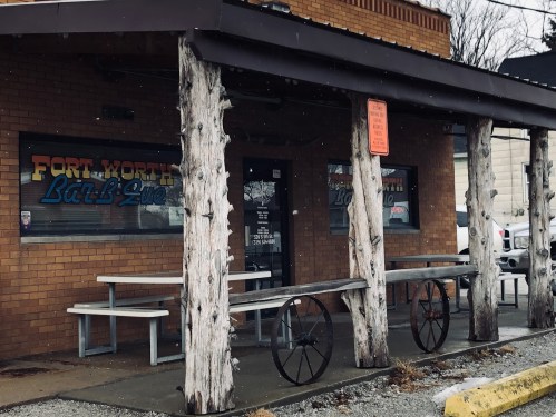 Exterior of a rustic building with wooden posts, a sign reading "Fort Worth BBQ," and picnic tables outside. Snow is falling.