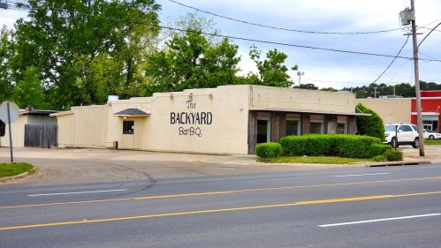A roadside view of "The Backyard Bar-B-Q," a single-story building with greenery and a parking area.