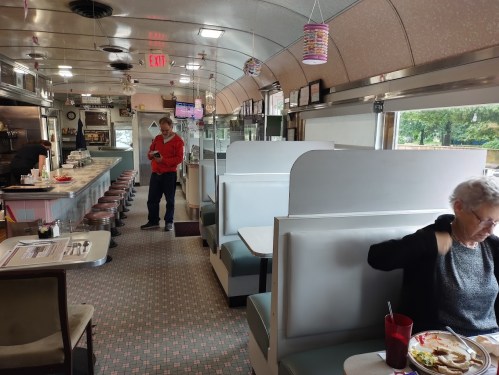 A retro diner interior with booths, a counter, and a woman eating while a man stands nearby, looking at his phone.