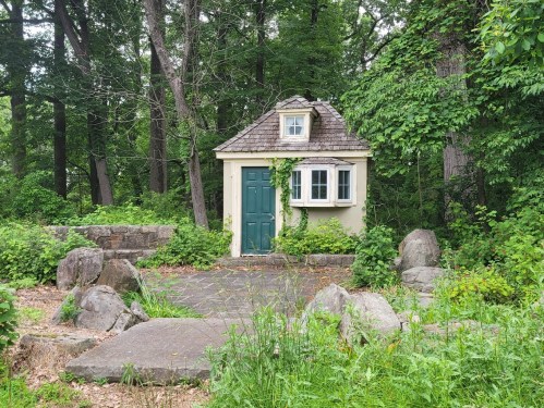 A small, charming house with a green door, surrounded by lush greenery and large rocks in a wooded area.