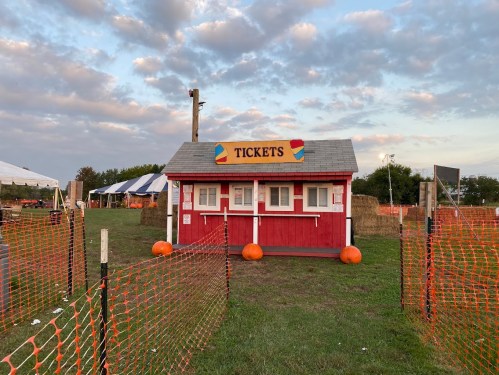A red ticket booth with a sign reading "TICKETS," surrounded by pumpkins and orange fencing in an outdoor setting.