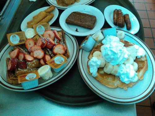 A tray with two plates of breakfast: one with French toast topped with strawberries and syrup, the other with waffles and whipped cream.