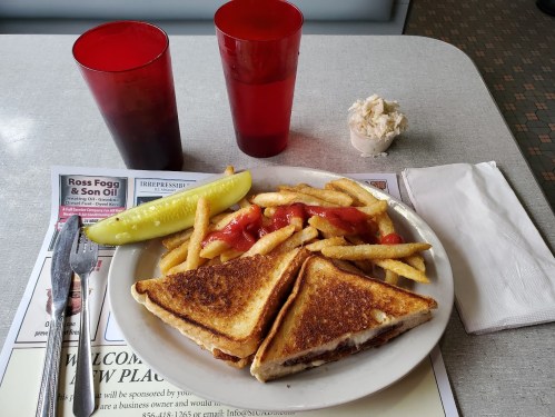 A plate with a grilled cheese sandwich, fries, a pickle, and ketchup, accompanied by two red cups and a small dessert.