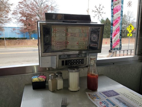 A vintage jukebox sits on a diner table, next to a bottle of ketchup and condiments, with a view of the street outside.