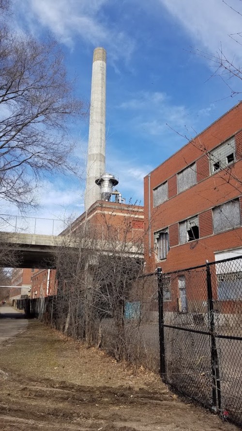 Abandoned industrial building with a tall smokestack, surrounded by bare trees and a chain-link fence under a blue sky.