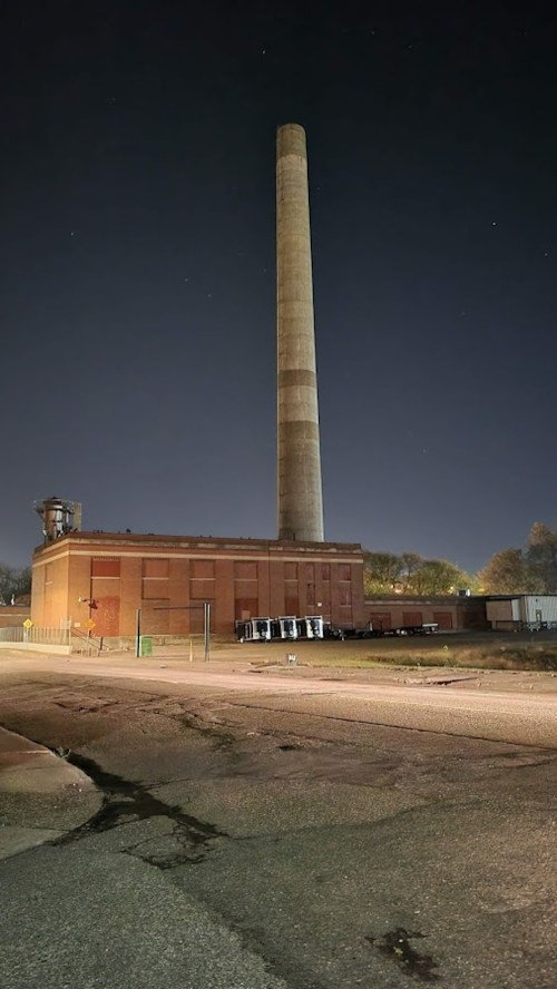 A tall smokestack rises from a brick industrial building at night, with a clear sky and distant lights in the background.