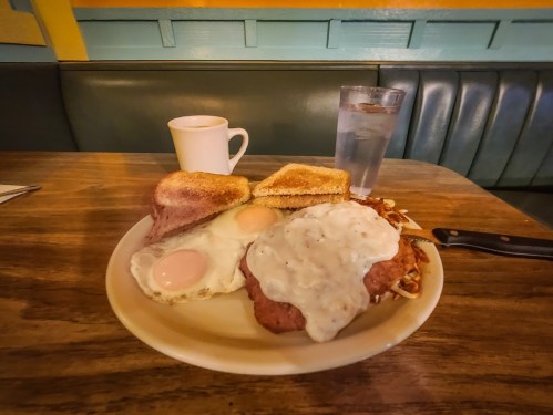 A plate of breakfast featuring two eggs, chicken fried steak with gravy, toast, and a cup of coffee beside a glass of water.