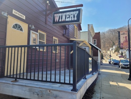 A wooden winery building with a sign, featuring a ramp and entrance, set in a quaint street scene.