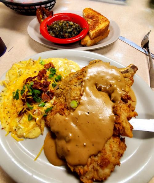 A plate of fried chicken with gravy, served with cheesy scrambled eggs, bacon, and a side of collard greens and toast.