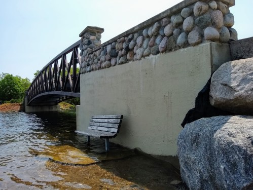 A wooden bench sits partially submerged in water near a stone wall and a bridge under a clear blue sky.