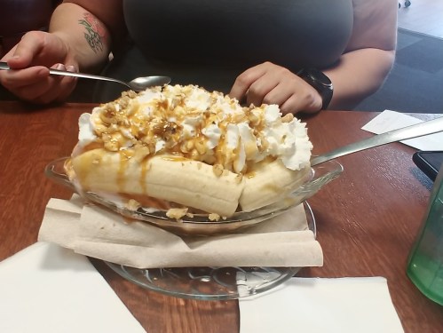 A glass dish filled with bananas, whipped cream, caramel, and popcorn, with a spoon resting beside it.