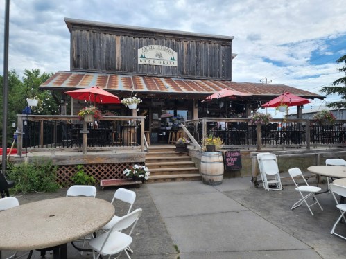 A rustic bar and grill with a wooden exterior, outdoor seating, and red umbrellas on a cloudy day.