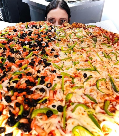 A person peeks over a large pizza divided into two halves, one with vegetables and the other with black olives and toppings.