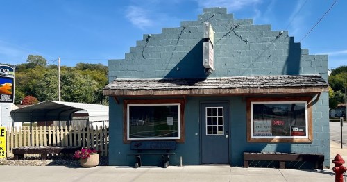 A blue, triangular-roofed building with a wooden porch, flower pots, and a sign indicating it's open.