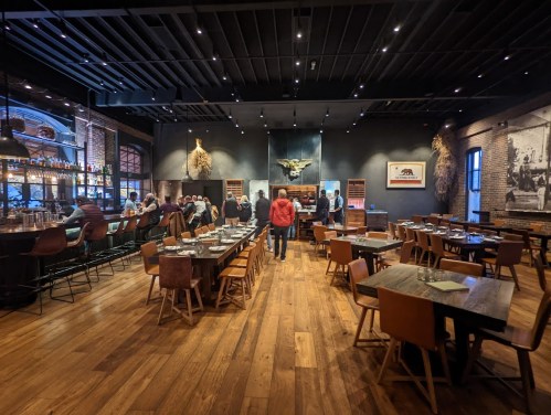 A spacious restaurant interior with wooden floors, tables, and a bar, featuring diners and a warm, inviting atmosphere.