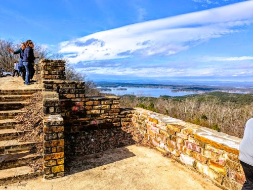 Two people stand on a stone overlook, enjoying a scenic view of a lake and mountains under a blue sky with clouds.