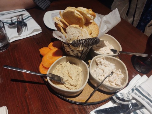 A basket of toasted bread slices surrounded by four bowls of spreads, including hummus and chicken salad, on a wooden table.
