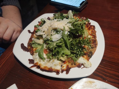 A white plate with a pizza topped with fresh greens, tomatoes, and cheese, resting on a wooden table.