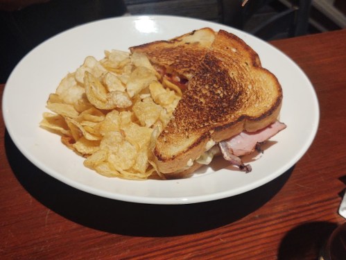 A toasted sandwich with ham and cheese, served with a side of crispy potato chips on a white plate.