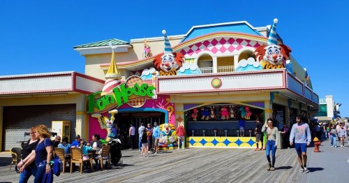 Colorful amusement arcade on a boardwalk, featuring clown decorations and people walking by. Bright blue sky above.