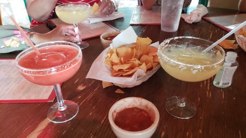 A table with two colorful cocktails, a basket of tortilla chips, and a small bowl of salsa.