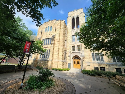A stone building with large windows, surrounded by trees and greenery, under a blue sky. Pathway leads to the entrance.