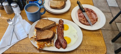 A breakfast plate with two eggs, bacon, hash browns, toast, and a side of ham, with a cup of coffee nearby.
