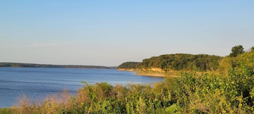 A serene lakeside view with lush greenery and a clear blue sky, showcasing gentle hills in the background.