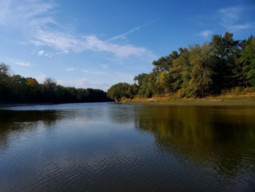 A serene river scene with calm water reflecting trees and a clear blue sky.