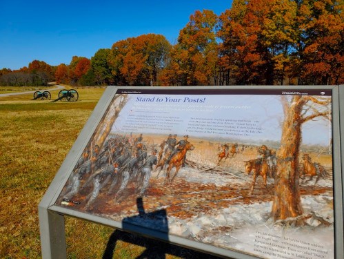 A historical marker with a painting of soldiers and cannons, set against a backdrop of autumn trees.