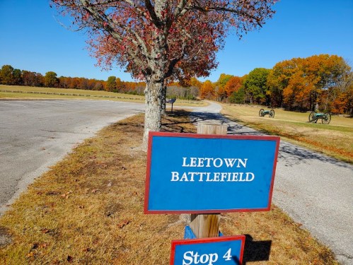 Sign for Leetown Battlefield with a tree and autumn foliage in the background, near a road and historical cannons.