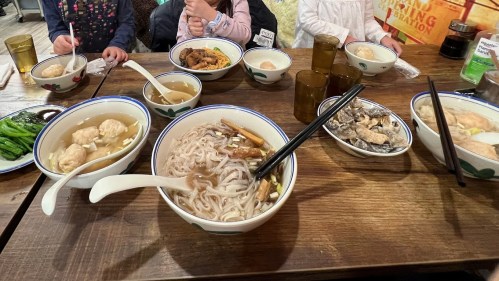 A table filled with various dishes, including noodles, soup, and side dishes, with people’s hands visible.