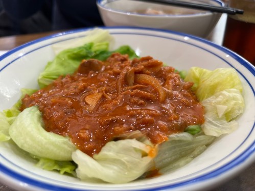 A bowl of lettuce topped with a savory red sauce and sliced ingredients, served alongside a bowl of soup.