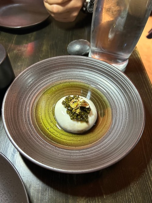 A small dish featuring a creamy white sphere topped with green sauce and garnished, placed on a textured plate.
