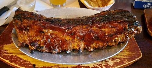 A large, glazed rack of ribs on a metal platter, served with a side of sauce and garnished with a slice of lemon.