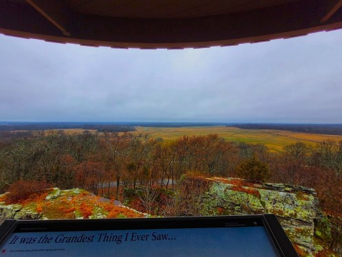 A scenic view from a lookout point, featuring a vast landscape of fields and trees under a cloudy sky.