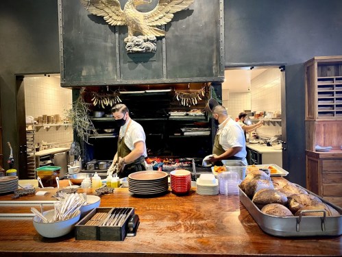 Two chefs work in a bustling kitchen, preparing food at a large wooden counter with various dishes and utensils.