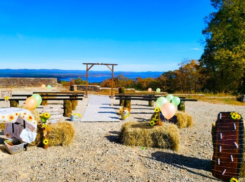 Outdoor wedding setup with hay bales, wooden benches, balloons, and sunflowers against a scenic mountain backdrop.