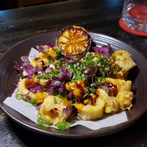 A plate of crispy calamari topped with herbs, drizzled with sauce, and garnished with a grilled lemon slice.