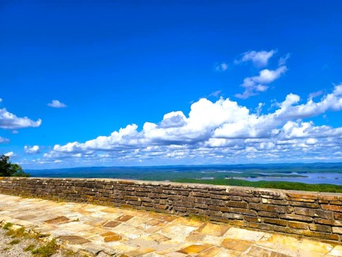 A scenic view from a stone wall overlooking a lake and rolling hills under a bright blue sky with fluffy clouds.
